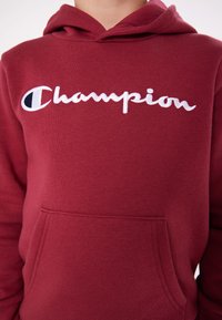 Felpa con cappuccio bordeaux in tessuto morbido, dotata di una tasca davanti e di un logo "Champion" ricamato in bianco con un accento blu sulla lettera "C".