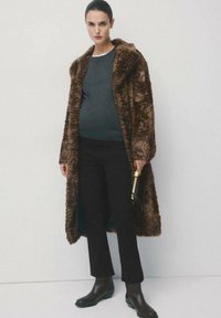 Kobieta w ciąży ubrana w brązowy futrzany płaszcz w wzory, ciemnozieloną bluzkę, czarne spodnie i brązowe botki, trzymająca małą, złoto-czarną kopertówkę.