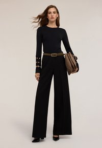 Femeie îmbrăcată într-un top negru cu mâneci lungi și tăieturi, pantaloni negri evazați, curea cu cone, pantofi cu toc negri și ținând o poșetă maro.