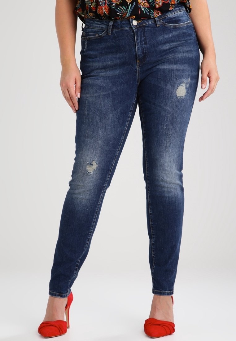 Jean en denim bleu foncé coupe slim, avec des détails usés aux genoux et une taille haute. Associé à des chaussures à talons hauts rouges.