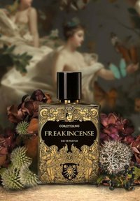 Bottiglia di profumo in vetro nero con ornate rifiniture dorate, caratterizzata da cherubini e un motivo decorativo, etichettata "Freakincense" su uno sfondo floreale.