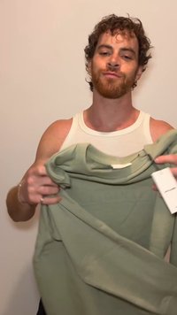 Hombre con cabello rizado y barba sostiene una sudadera verde clara con una etiqueta visible, de pie frente a una pared beige lisa.