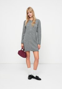 Γκρι πλεκτό φόρεμα με μακριά μανίκια, κουμπιά κατά μήκος του μπροστινού μέρους και ίσια γραμμή. Συνδυασμένο με λευκές κάλτσες και μαύρα παπούτσια.