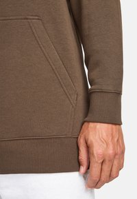Brauner Sweatshirt-Stoff mit Kängurutasche, gerippten Bündchen und Saum. Verfügt über eine glatte Textur und eine taillierte Passform an den Handgelenken.