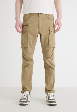Kahaki cargo pants with multiple pockets and zippers, featuring a relaxed fit, stitched detailing, and durable fabric, paired with sneakers.