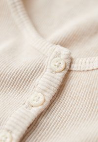 Beige ribbet stof med en knapstolpe, der har hvide knapper. Nærbillede, der fremhæver tekstur og fine detaljer i stoffet.