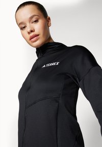 Čierna zip-up bunda TERREX s vysokým golierom, hladkou textúrou a jemnými švovými detailmi, navrhnutá na vonkajšie aktivity.