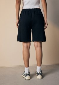 Schwarze Baumwollshorts mit ausgefranstem Saum, elastischem Bund und Seitentaschen, kombiniert mit hellen Sneakers und weißen Sportsocken.