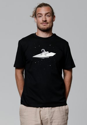 T-shirt en coton noir avec un motif d'astronaute blanc sur un avion en papier, le tout sur fond étoilé. Manches courtes et coupe décontractée.