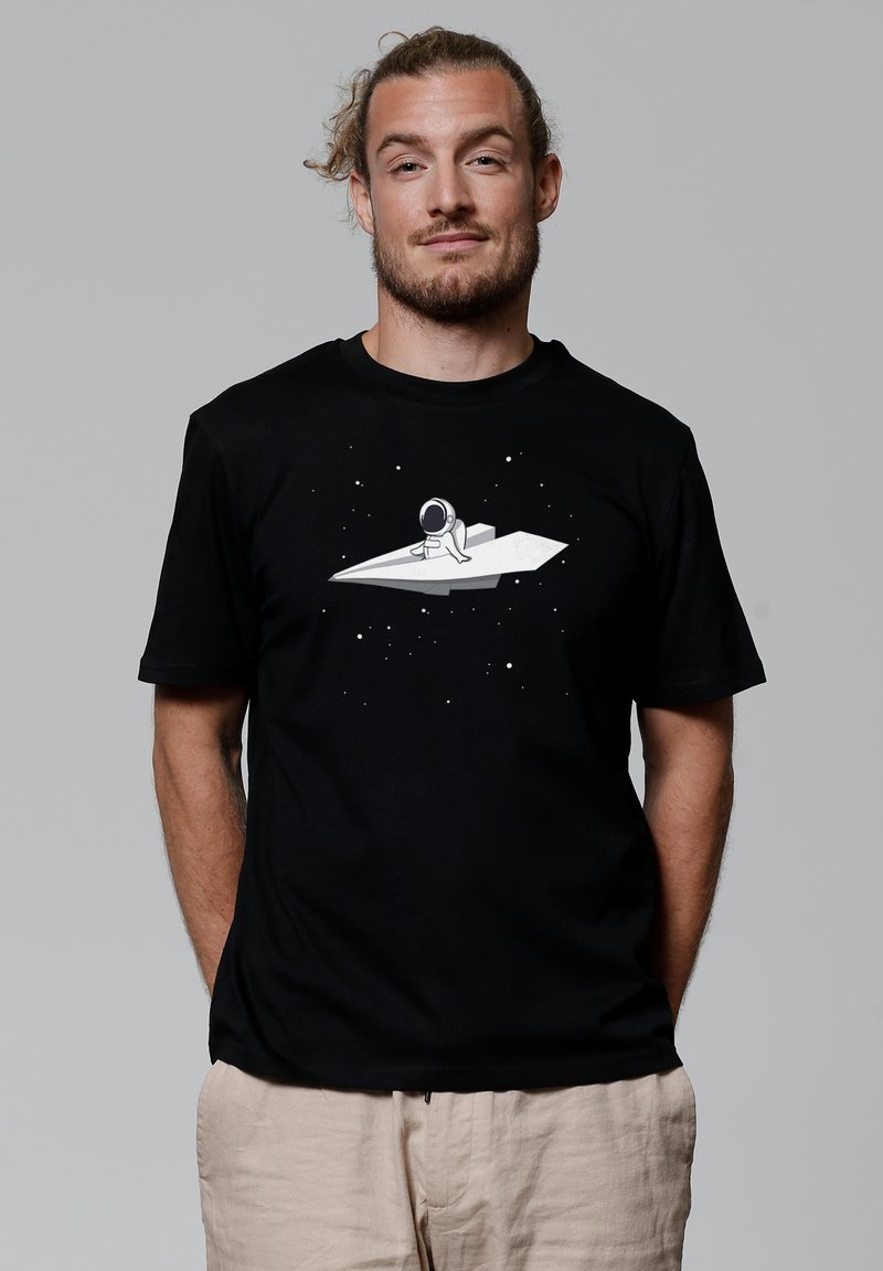 Zwarte katoenen t-shirt met een witte astronautafbeelding op een papieren vliegtuigje, tegen een sterrenachtergrond. Korte mouwen en een ontspannen pasvorm.