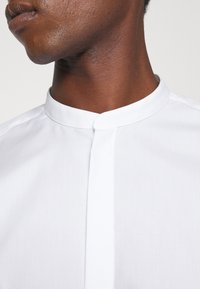 Gros plan d'une personne portant une chemise blanche à col mandarin, montrant le cou et la partie inférieure du visage.