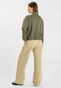 Pull olive avec col montant et ourlet élastique, associé à un pantalon beige à jambes larges avec poches arrière et tissu texturé.