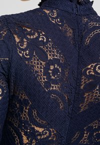 Tissu en dentelle bleu marine présentant des motifs floraux et géométriques complexes avec des accents texturés. Le design a une apparence superposée.