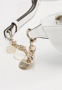 Valkoinen keinonahkainen käsilaukku hihna ruskeilla yksityiskohdilla. Kultaista metalliosaa ja kolme pyöreää riipusta, joissa on kaiverrettuna "GUESS EST. 1981". Rakeinen pinta.