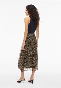 Kvinne med krøllete hår iført en svart ermeløs topp, brun og svart blomstret midiskjørt, og svarte hælesandaler, som står i profil.