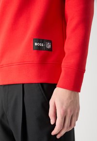 Sweatshirt vermelho feito de mistura de algodão, com uma etiqueta preta BOSS x NFL na bainha. Punhos padrão e silhueta reta. Calças pretas visíveis.