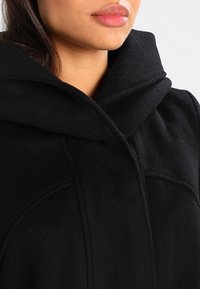 Svart huva kappa med en mjuk textur och en skräddarsydd passform. Har en hög krage och subtil sömnadsdetaljering för en ren, modern look.