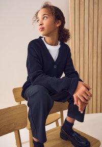 Temen pleteni kardigan z globokim V-izrezom, dvema gumboma, kombiniran s črnimi hlačami in sijajnimi mokasini. Kodrasti lasje in mehka osvetlitev poudarjata videz.