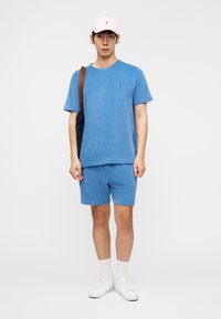 Νέος άνδρας που φοράει μπλε μπλουζάκι και σορτς, ροζ καπέλο, λευκά αθλητικά παπούτσια και κάλτσες, κρατώντας μαύρη τσάντα ώμου με καφέ λουράκι.