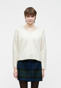 Cremefarget cardigan med V-hals, lange ermer og fem knapper foran. Teksturert, myk stoff med cropped lengde. Kombinert med rutete skjørt.