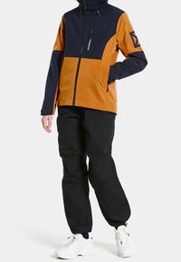 Color-blocked waterproof jacket in orange and navy, featuring a zippered front, hood, and angled pocket, paired with black pants and white sneakers.
