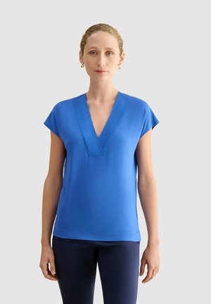 Mujer de piel clara y cabello rubio que lleva pendientes dorados, una blusa azul de manga corta con cuello en V y pantalones oscuros sobre un fondo liso.