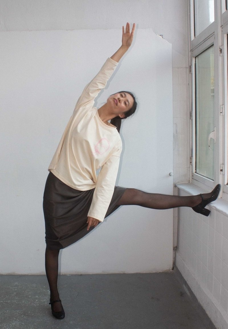 Krémová košile s dlouhým rukávem a růžovou grafikou, hnědá saténová sukně, černé punčochy a třpytivé lodičky, pózující u okna.