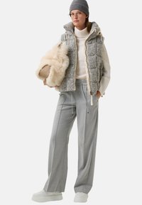 Szary pikowany kamizelka z wysokim kołnierzem, biały sweter z dzianiny, szare spodnie i masywne białe trampki. Trzyma miękką beżową torbę.