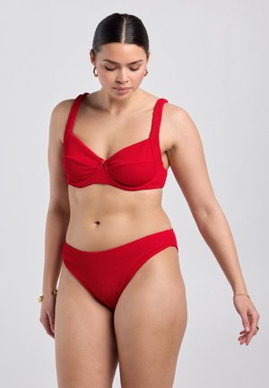 Frau, die einen strukturierten roten Bikini mit breiten Trägern trägt, steht vor einem einfachen hellgrauen Hintergrund und schaut nach unten und zur Seite.