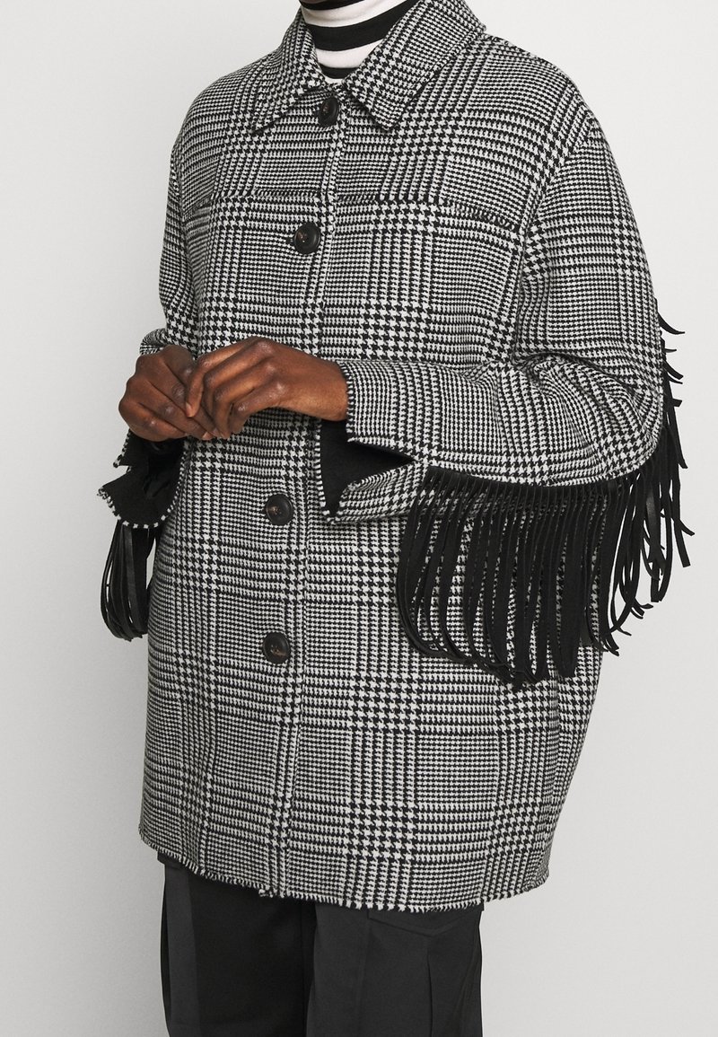 Abrigo de cuadros en blanco y negro con detalles de flecos en las mangas, con cuello, dos botones y un corte holgado. Textura de tela tejida suave.
