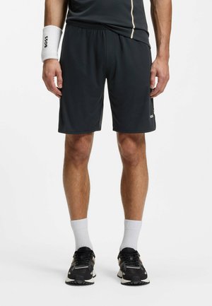 Homme portant un short de sport noir, des baskets noires, des chaussettes blanches montantes, un haut noir avec un liseré beige, et un bracelet blanc.
