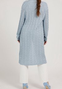 Cárdigan largo de punto azul claro con patrones de trenzas, de mangas largas y con laterales abiertos, combinado con pantalones blancos y zapatos de tacón azules.