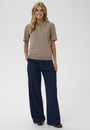 Beige Poloshirt mit kurzen Ärmeln und drei Knöpfen, kombiniert mit weiten dunklen Denim-Hosen, Modell steht vor einem neutralen Hintergrund.