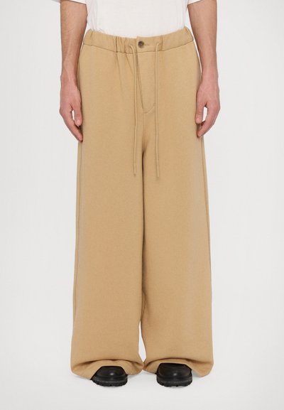 Beige weit geschnittene Sweatpants mit einem Kordelzugbund, ausgestattet mit einer weichen Textur und einer vorderen Tasche, kombiniert mit schwarzem Schuhwerk.