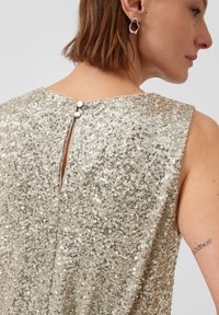 Zbliżenie kobiety w sukience bez rękawów z srebrnych cekinów z detalem guzika na plecach oraz nieregularnymi srebrnymi kolczykami.