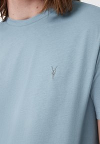 T-shirt in cotone azzurro chiaro con collo rotondo. Presenta un piccolo logo ricamato di teschio di capra sul lato sinistro del petto. Texture morbida, design casual.