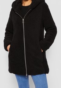 Abrigo de forro polar negro con capucha, cierre frontal completo, dos bolsillos laterales y un acabado suave y texturizado. Combinado con unos jeans de denim azul.