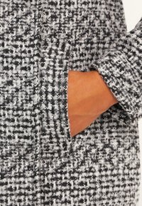 Černobílý tvídový kabát s texturovaným povrchem, který má postranní kapsu a moderní střih, s rukou položenou uvnitř kapsy.