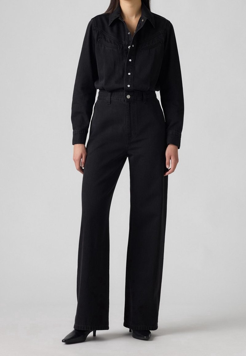 Femme portant une chemise noire boutonnée rentrée dans un pantalon noir taille haute à jambes larges avec des talons noirs pointus, sur un fond uni.