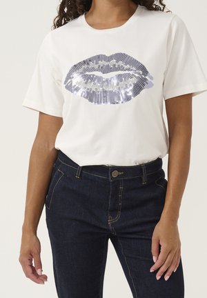 Femme portant un T-shirt blanc à manches courtes avec un design de lèvres en sequins argentés, associé à un jean bleu foncé, se tenant avec les bras détendus.