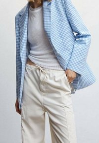 Blazer a quadri azzurro chiaro, canotta bianca a coste e pantaloni beige con cordoncino. Il blazer presenta due bottoni e una tasca frontale.