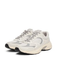 Chaussures de sport beiges et grises avec des panneaux en maille, des superpositions texturées et une pointe arrondie. Elles possèdent des lacets plats et une semelle en caoutchouc noire.