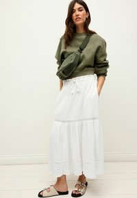 Sweatshirt vert court, jupe maxi blanche à volants avec un tissu texturé et des détails en dentelle, et sandales à glisser beiges avec des accents foncés.