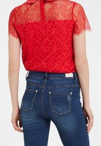 Blusa de encaje rojo con cuello de Peter Pan, combinada con jeans ajustados de un azul oscuro. La blusa presenta un diseño estampado y mangas transparentes.