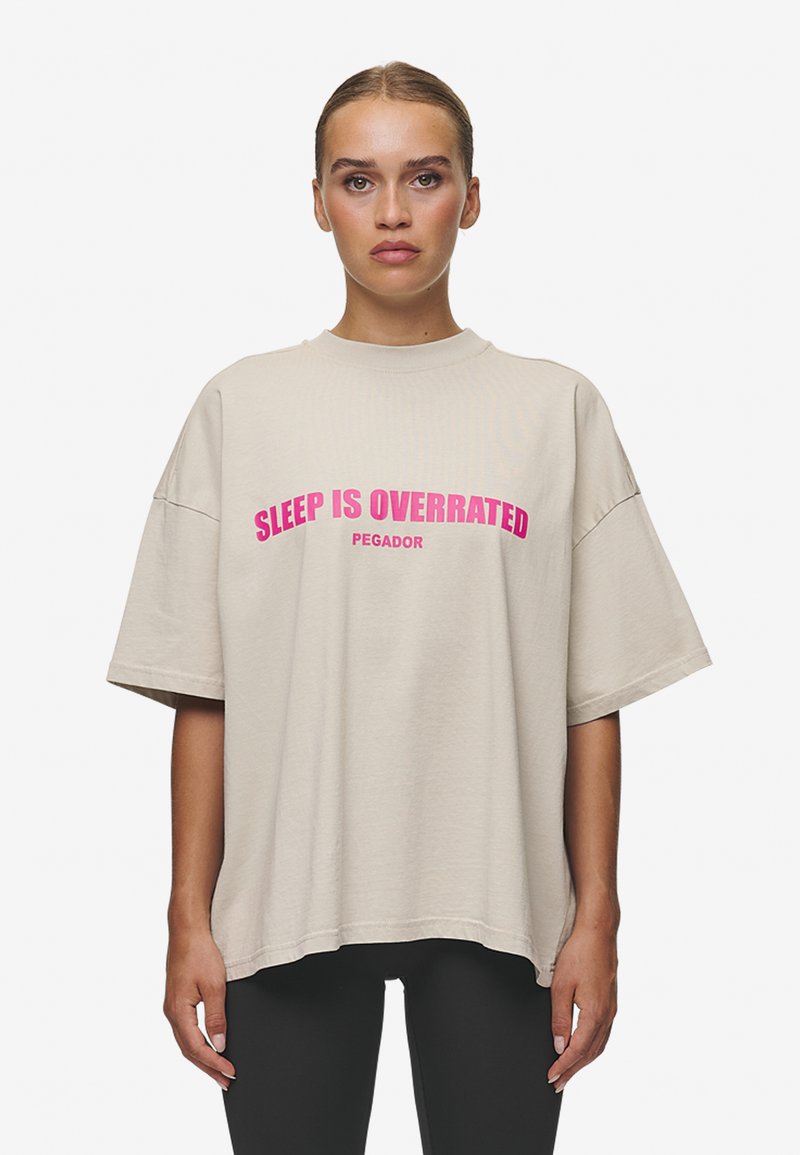 Übergroßes, beiges T-Shirt aus Baumwolle. Mit auffälligem pinkem Text: "SCHLAF IST ÜBERBEWERTET" mit "PEGADOR" darunter. Kurze Ärmel und entspannter Schnitt.