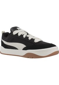 Scarpa sportiva in bianco e nero con texture liscia e scamosciata, punta tondeggiante, suola beige e logo prominente sulla linguetta. Presenta lacci bianchi.