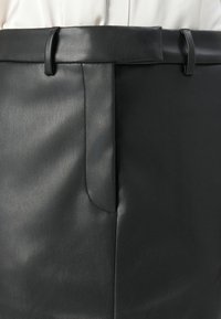 Gros plan sur une jupe en cuir noire taille haute avec des passants de ceinture, une taille repliée et une fente à l'avant, portée sur un haut blanc.