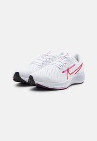 Une paire de chaussures de course Nike blanches avec un swoosh rose, une tige en mesh et des semelles noires et violettes, angulées sur un fond blanc.