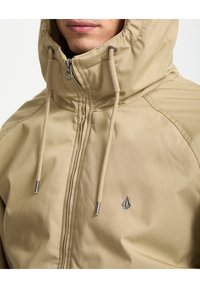 Giacca beige con cappuccio, cerniera lampo, coulisse e logo sul petto. Presenta una texture liscia e opaca e un design strutturato.