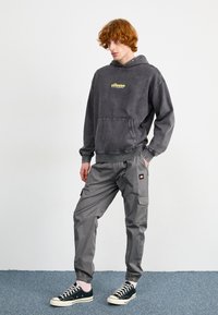 Hoodie gris délavé avec une poche frontale et un logo jaune, associé à un pantalon cargo gris avec des poches latérales et des poignets élastiques. Baskets noires.